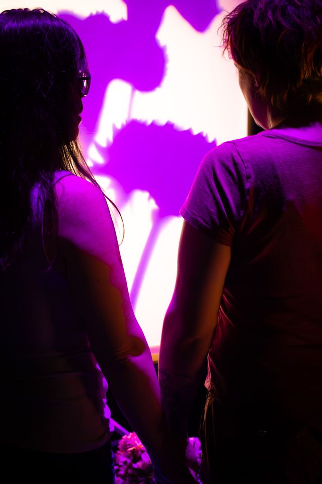 Two people from behind holding hands and a bundle of flowers between them, looking at a screen with projected flower shadows in purple