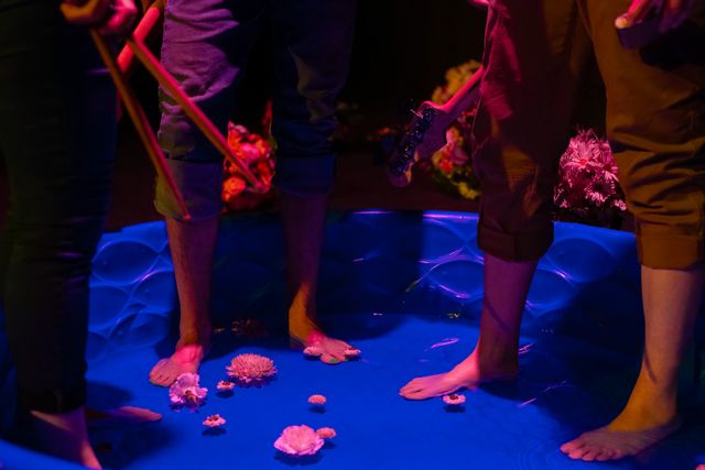 Three pair of feet with rolled-up trousers standing in a kiddie pool with floating flowers, one hand holding drum sticks, guitars slung on backs, and flowers in  the background