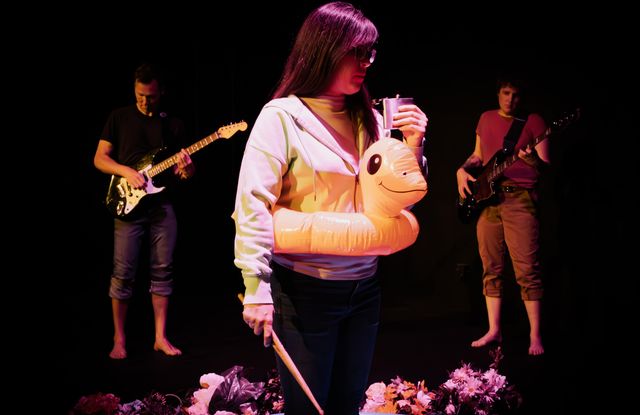 Sondra standing in a kiddie pool, with drum sticks and a flask of whiskey, and a duck floaty around her waist - Dan and Mia in the back barefoot playing guitar and bass on a black background - everyone looks kinda sad