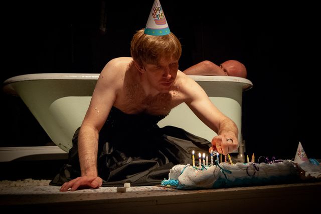 Mat Shultz in a black dress & birthday hat, lighting candles on a cake - in front of a bathtub
