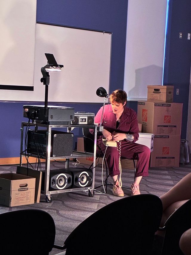 Miriam sits next to an AV cart with an overhead projector, surrounded by cardboard boxes