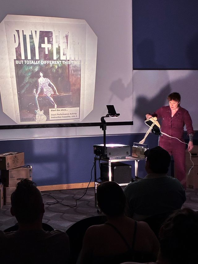 Miriam in a boiler suit setting a landline phone next to an overhead projector showing the pity+fear title slide