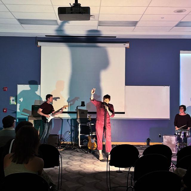 Dan playing guitar, Miriam with a mic gesturing (her shadow large behind her), and Sondra playing a drum kit with an old suitcase as the kick drum