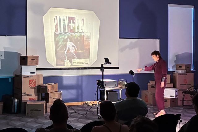 Miriam is pointing at stacked boxes. There's an AV cart in the center of the space with an overhead projector showing a title slide for the show.
