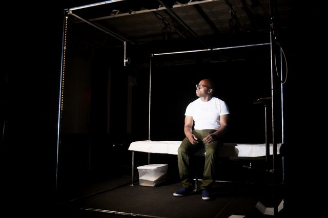 a Black man sits on a plain cot wearing prison greens, in a six foot cube made of bare pipe that seems to glow from the inside